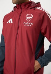 Röd och marinblå vindjacka med dragkedja, huva, broderad Arsenal-logga och vit sponsortext. Har Adidas-logotyp och elastiska ärmar.