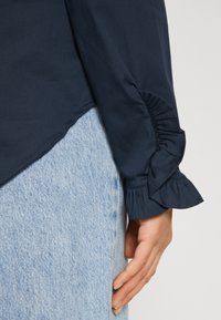 Blouse bleu marine avec des poignets à volants et un ourlet légèrement courbé, associée à un denim bleu clair. Tissu lisse, coupe ajustée et éclat subtil.