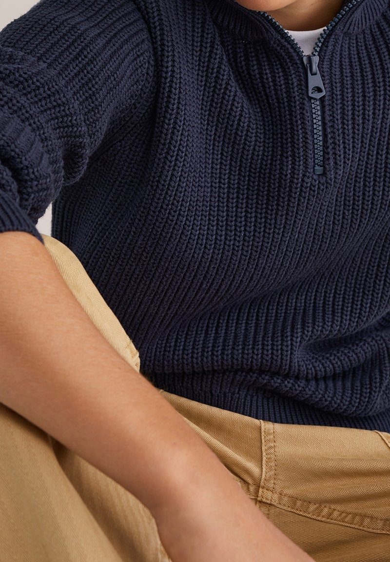 Pull en maille côtelée bleu marine avec col court zippé porté avec un pantalon beige, montrant le bras et le torse dans une pose détendue.