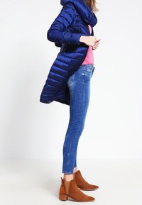 Marineblauwe gewatteerde jas met capuchon, roze top, blauwe skinny jeans en bruine suède enkellaarsjes met een lage hak en uitsnijding.