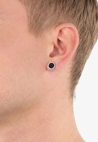 Pendiente de botón negro con un delgado borde exterior plateado, forma circular, textura lisa, llevado en una oreja perforada con tono de piel claro y cabello corto.