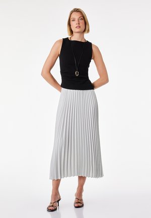 Donna che indossa un top nero senza maniche, gonna lunga bianca plissettata, sandali neri con tacco e collana con pendente, in piedi con le mani dietro la schiena.