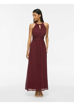 Femeie purtând o rochie maxi burgundy fără mâneci, cu detalii din dantelă în talie și pantofi cu toc deschis în față, stând într-un fundal alb simplu.