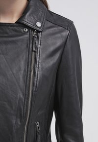 Close-up of a black leather jacket with silver zipper and snap button collar worn on a person’s upper body and arm.