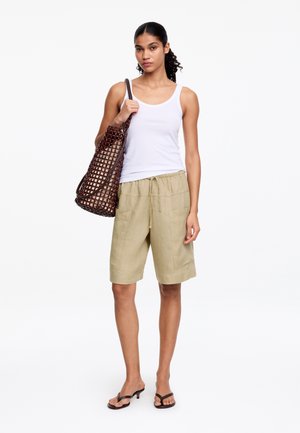 Frau trägt weißes Tanktop, beige knielange Shorts, schwarze Sandalen mit Absatz und trägt eine große braune gewebte Tragetasche über der Schulter.