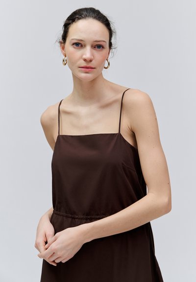 Jeune femme aux cheveux foncés portant des boucles d'oreilles créoles dorées et une robe brune sans manches, debout devant un fond clair uni.