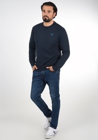 Navy sweatshirt with round neckline, ribbed cuffs, and casual fit, paired with blue jeans and white sneakers. Features a small blue logo.