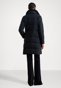 Tamsiai mėlynas siuvinėtas pūkinis paltas su aukštu apykakle, ilgomis rankovėmis ir vidutinio ilgio kirpimu, derinamas su tamsiomis plačiomis kelnėmis.