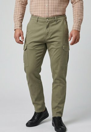 Mann trägt olivgrüne Cargohosen, schwarze Sneakers und ein beiges kariertes Hemd, steht vor einem einfarbigen Hintergrund.