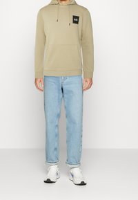 Sudadera beige con un bolsillo delantero, capucha con cordón y parche de logo negro, combinada con jeans azul claros y zapatillas deportivas blancas.