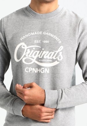 Person iført en grå sweatshirt med hvid tekst: "Handmade Garments EST. 1990 Originals by Jack & Jones CPNHGN."