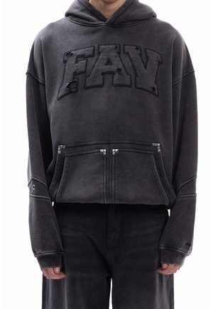 Personne portant un sweat à capuche noir avec le texte « FAV » et des détails usés, poche avant avec des clous en métal, associé à un pantalon noir.