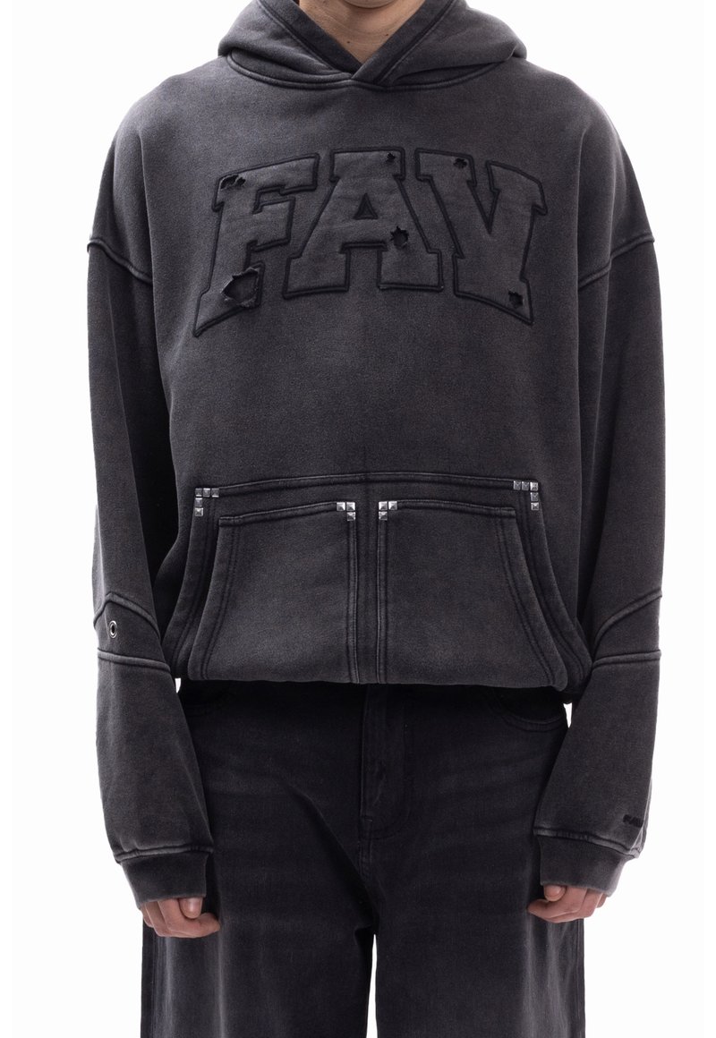Personne portant un sweat à capuche noir avec le texte « FAV » et des détails usés, poche avant avec des clous en métal, associé à un pantalon noir.