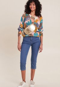 Pull en tricot coloré avec des motifs géométriques, associé à un jean bleu court et une banane dorée brillante. Des baskets blanches viennent compléter le look.