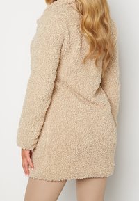 Beige, fluffy fleecejakke med en struktureret overflade, lange ærmer og en krave, der ender over knæet, med en afslappet pasform.