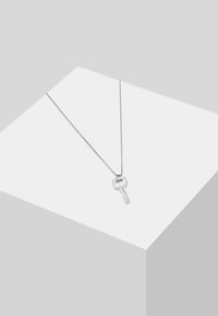 Collana con pendente a forma di chiave in argento su una fine catena. La chiave presenta una testa arrotondata e scanalature. Design minimalista su uno sfondo grigio chiaro.