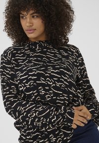 Un haut à manches longues avec un col montant, présentant un motif de vagues noir et beige, fabriqué en tissu texturé. Les manches à poignet complètent le design.