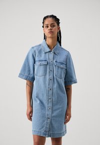 Lys blå denimkjole med korte ermer, to brystlommer og knappelukking foran. Glatt tekstur og rett snitt.