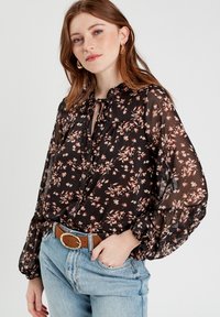 Blouse noire à fleurs avec des manches longues transparentes, des poignets froncés et un col à nouer. Présente des motifs de petites fleurs roses et blanches. Portée avec un jean bleu.