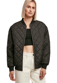 Schwarze gesteppte Bomberjacke mit gerippten Bündchen und Kragen, ausgestattet mit einem Frontreißverschluss und einem rautenförmigen Strukturmuster. Getragen über einem schwarzen Crop-Top.