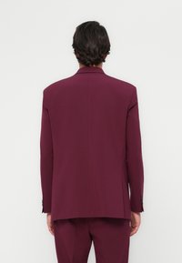 Giacca da abito bordeaux con silhouette strutturata, spacco singolo e maniche lunghe. Presenta polsini con bottone e una texture di stoffa liscia.