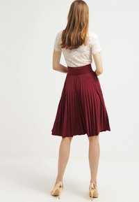 Donna con capelli lunghi che indossa un top in pizzo bianco, una gonna plissettata bordeaux e scarpe con tacco dorate, in piedi con le mani sui fianchi e rivolta di spalle.
