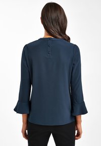 Námořnická modrá blůza s tříčtvrtečními rukávy, rozšířenými manžetami a třemi knoflíky na zadním výstřihu. Hladká, lehká tkanina.