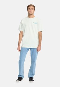 Lichtgroen katoenen t-shirt met korte mouwen en een turquoise "SPYDER" logo op de borst, gecombineerd met lichtblauwe denim jeans en zwarte sneakers.
