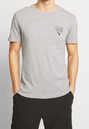 Homme portant un t-shirt gris clair avec un petit motif noir de montagne et soleil sur la poitrine gauche et un pantalon noir, les mains dans les poches.