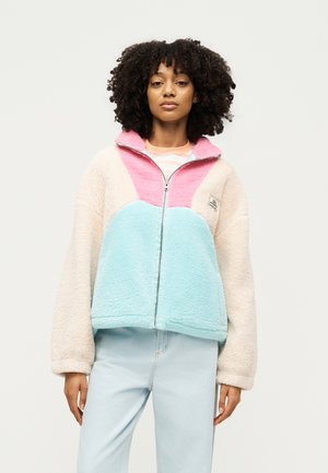 Kvinde med krøllet hår iført en pastelrosa, blå og beige fleecejakke med lynlås og lyseblå jeans, stående foran en ensfarvet baggrund.