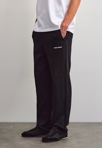 Person trägt schwarze Pegador-Sweathosen und schwarze Sneakers, hat die Hände in den Taschen und steht auf grauem Boden vor einer weißen Wand.