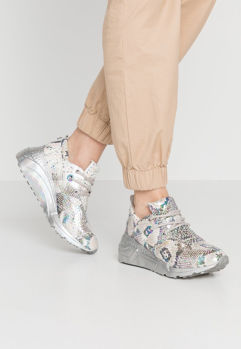 Personne portant un pantalon de jogging beige et des baskets épaisses argentées brillantes à motif peau de serpent, debout sur un sol clair et lisse.