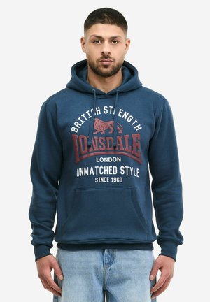 Muž v modré mikině Lonsdale s nápisem "British Strength", lvím logem a textem "Unmatched Style Since 1960", doplněný světlými modrými džínami.