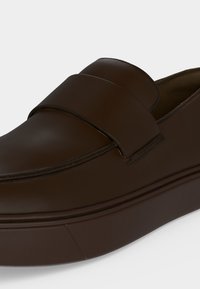 Σκούρο καφέ παπούτσι slip-on με λείο δέρμα στην επάνω επιφάνεια και παχιά λαστιχένια σόλα, που εμφανίζει λεπτομέρειες από πλευρά και πάνω φόντο λευκό.