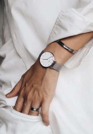 Main portant une montre en maille argentée, un bracelet argenté et une bague argentée, reposant sur un tissu blanc avec la main partiellement dans une poche.