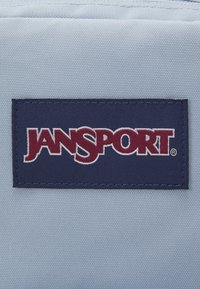 Light blue fabric background with a navy rectangular patch displaying the "Jansport" logo in burgundy, featuring white outlining.