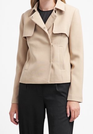 Žena v béžovém saka na míru s překládanými předními panely a detaily se stiskacími knoflíky, v kombinaci s černými kalhotami.