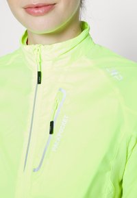 Veste jaune néon avec un col haut, une fermeture éclair à l'avant et le texte "PACKPOCKET" ; présente un tissu léger et texturé ainsi que des accents réfléchissants.