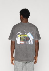 Γκρι βαμβακερό t-shirt με χαλαρή εφαρμογή, που διαθέτει εκτυπωμένα δελτία επιβίβασης και λεπτομέρειες αναχώρησης αεροδρομίου στην πλάτη, περιβαλλόμενο από μοτίβα φυσαλίδων.