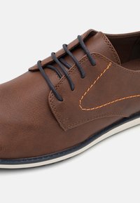 Zapato de cuero marrón con una superficie texturizada, con acentos de costuras naranjas y cordones negros. La suela es blanca con un borde azul marino.