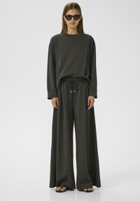 Ensemble composé d'un sweat-shirt gris oversize et d'un pantalon large, confectionné dans un tissu doux. Il présente une taille à cordon de serrage et des poignets côtelés, accompagné de sandales plates.