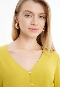 Blusa gialla con scollatura a V e tre piccoli bottoni sul fronte. Tessuto liscio, messo in evidenza da grandi orecchini a cerchio dorati.