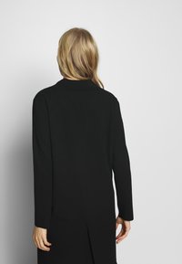 Svart långärmad kappa med mjuk textur, med skalkrage, minimalistisk design och ett slits bak för enkel rörelse.
