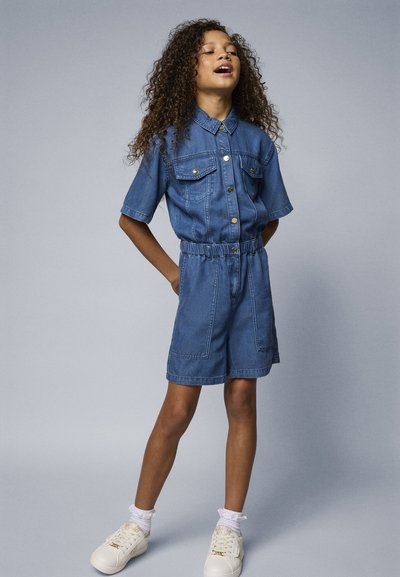 Mono de mezclilla en azul claro, manga corta, dos bolsillos en el pecho, cintura elástica y pantalones cortos adjuntos. Zapatillas blancas con cordones texturizados.