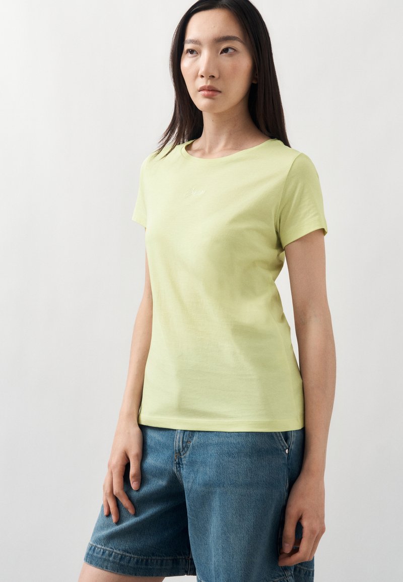 Γυναίκα με μακριά σκουρόχρωμα μαλλιά φοράει ένα ανοιχτοπράσινο κοντομάνικο μπλουζάκι και μπλε τζιν σορτς, στέκεται μπροστά σε ένα απλό λευκό φόντο.