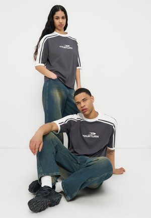 Bărbat tânăr așezat și femeie în picioare, amândoi purtând tricouri sportive negre și albe "Yourturn" și blugi albaștri deschiși, pe fundal alb.