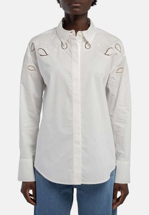Femme portant une chemise blanche à manches longues boutonnée avec une broderie cachemire sur les épaules, associée à un jean bleu.