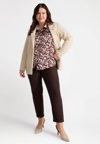 Blazer beige su misura sopra una camicia a righe marroni e bianche, abbinato a pantaloni marroni e tacchi beige chiari. Tessuto liscio e design aderente.