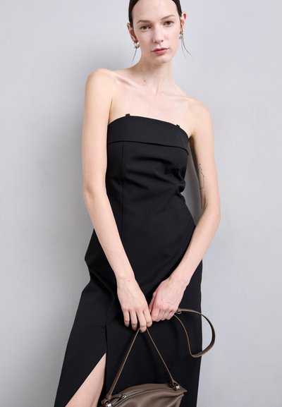 Black strapless dress in sleek fabric, featuring a fitted silhouette and side slit. Model holds a brown handbag. Simple, modern design.
