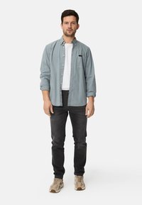 Light blue button-up shirt over a white t-shirt, paired with black jeans and beige sneakers. Shirt features a chest pocket and rolled sleeves.